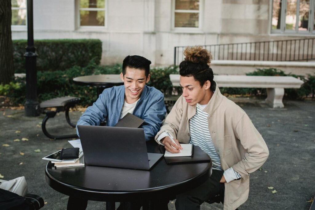 Smiling multiracial male classmates sitting at table and browsing netbook while taking notes in notebook